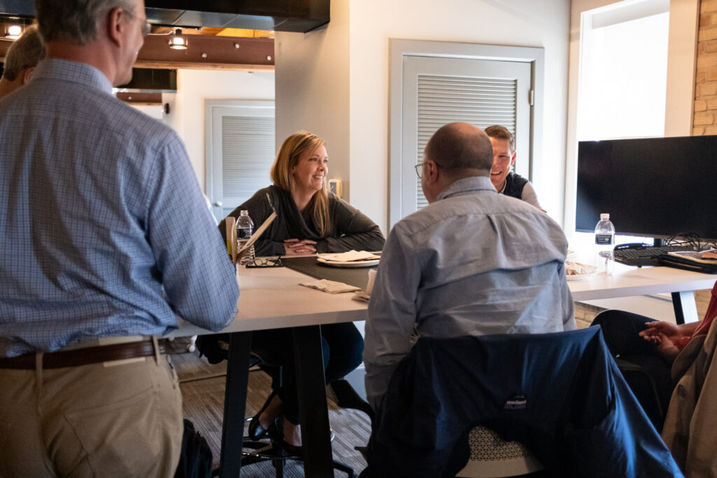 HDJ employees talking with one another. Two employees stand, while four employees sit around a long table with a computer at the end.