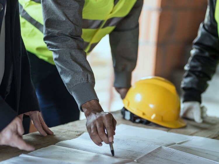 Construction workers lean over a set of blueprints, with one pointing at the paper with a pen.